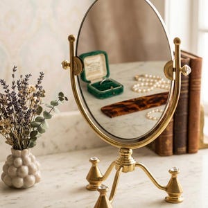 May include: A gold-toned oval tabletop mirror with a swivel frame supported by a three-legged stand. The base features small, clear, faceted crystal accents at the top of each leg and on the side adjustment knobs of the mirror frame.