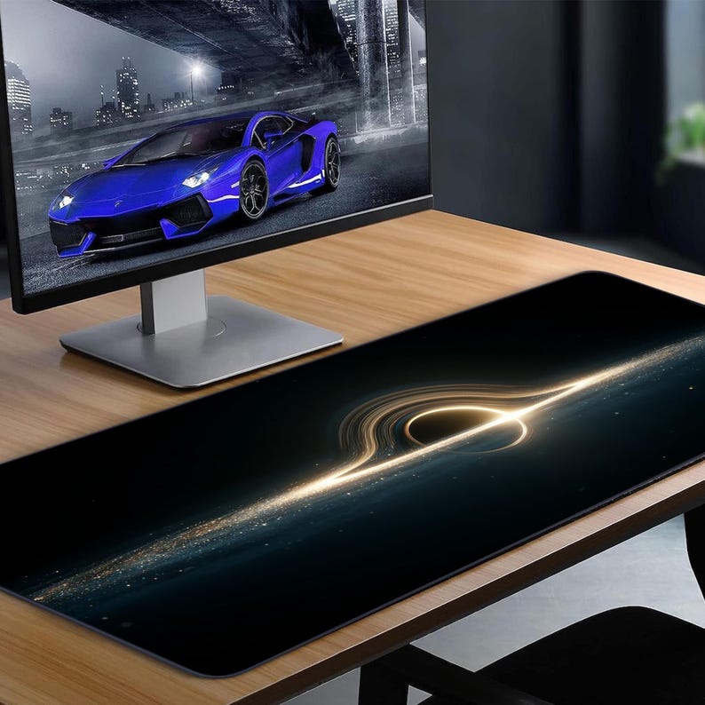 May include: Black desk mat featuring a glowing, golden spiral design. The mat is rectangular and placed on a wooden desk. A computer monitor shows a blue sports car on a city street.