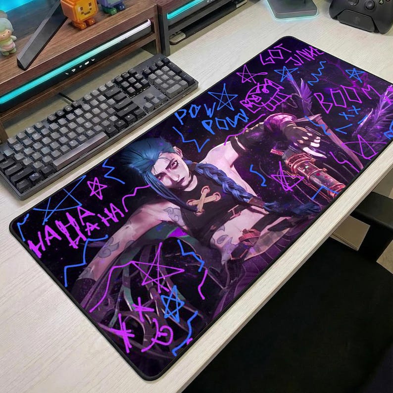 May include: A black rectangular mousepad featuring a graphic of a character with blue hair and a weapon, with the words "POW POW", "BOOM", and "HAHA" in neon colors. The mousepad is on a white desk next to a black keyboard.