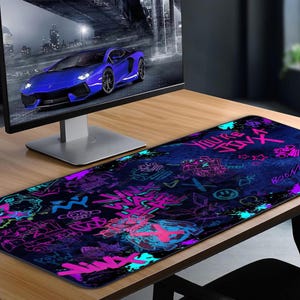 May include: A long, rectangular mouse pad with a dark blue background and graffiti-style art in neon pink, purple, and teal. The design includes text, symbols, and illustrations. A computer monitor and a blue sports car are in the background.