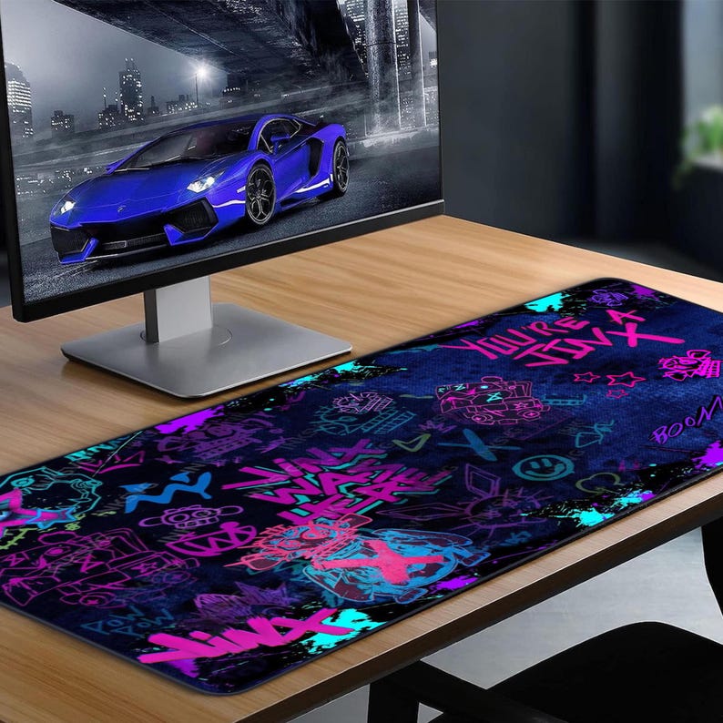 May include: A long, rectangular mouse pad with a dark blue background and graffiti-style art in neon pink, purple, and teal. The design includes text, symbols, and illustrations. A computer monitor and a blue sports car are in the background.