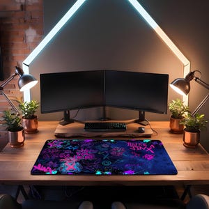 May include: A rectangular gaming mousepad with a vibrant design featuring neon pink and blue graphics on a dark blue background. The mousepad is placed on a wooden desk, with a keyboard, mouse, and dual monitors in the background.