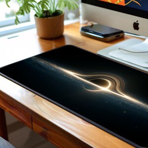May include: Rectangular black mousepad with a golden illustration of a black hole. The design shows a bright, circular centre and radiating light streaks against a dark backdrop. The mousepad is on a wooden desk.