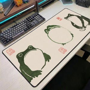 Puede incluir: Una alfombrilla de ratón rectangular con fondo color crema y borde negro. La alfombrilla presenta tres ilustraciones de ranas verdes al estilo artístico japonés, junto con sellos rojos. Un teclado negro es visible en el fondo.