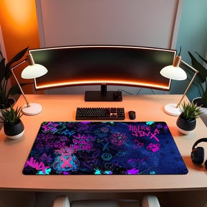 May include: A rectangular desk mouse pad with a dark blue background and graffiti-style artwork in neon pink, teal, and purple. The design includes text that reads "YOU'RE JINX" and other graphic elements. The pad is on a wooden desk with a keyboard, mouse, and monitor.