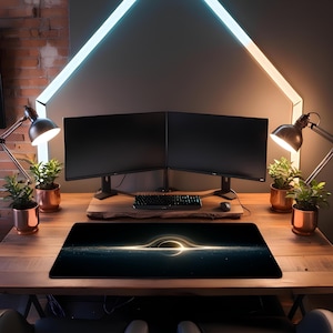 May include: A black desk mat with a glowing black hole design. The mat is rectangular and sits on a wooden desk, with a keyboard, mouse, and two computer monitors behind it. The desk is lit by desk lamps and a neon light fixture.