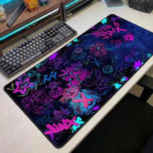 May include: A rectangular gaming mousepad with a dark blue background and neon pink and teal graffiti-style graphics. The design includes the words "You're Jinx" and "Jinx War". The mousepad is placed on a white desk next to a black keyboard.
