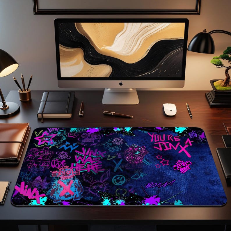May include: A rectangular mouse pad with a dark blue background and graffiti-style neon pink, purple, and teal graphics. The text "YOU'RE JINX" is visible. The pad is on a wooden desk with a computer and accessories.
