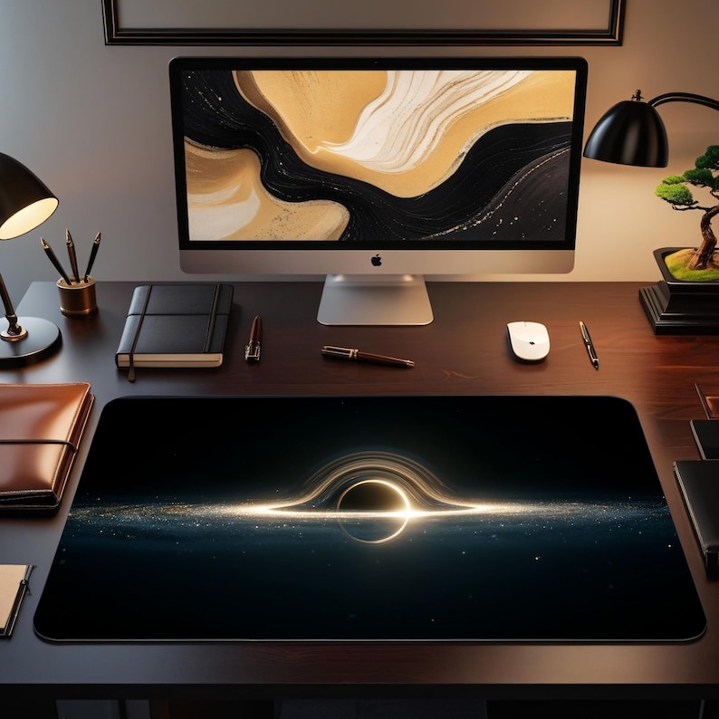 May include: A rectangular black mouse mat with a glowing black hole design. The image shows a dark, cosmic scene with a bright, circular light effect. The mouse mat is on a dark wooden desk with a computer and desk accessories.