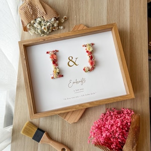 Könnte beinhalten: Ein rechteckiger Holzrahmen mit einem weißen Display, auf dem die Buchstaben I und J aus kleinen Trockenblumen in Rot-, Orange- und Gelbtönen geformt sind. In der Mitte befinden sich ein goldenes Et-Zeichen, der Text Embregts, 3 Juni 2022 und Ik hou van jou - Ich liebe dich.