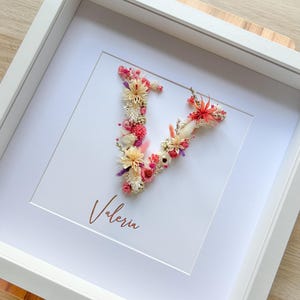 Könnte beinhalten: Gerahmtes Blumenkunstwerk mit dem Buchstaben "V", gefertigt aus getrockneten Blumen in Rosa-, Rot- und Cremetönen. Der Name "Valeria" ist darunter in Schreibschrift geschrieben. Das Kunstwerk ist in einem weißen Rahmen präsentiert.