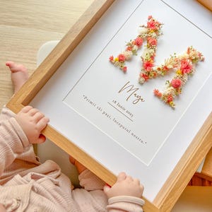 Pode incluir: Uma obra de arte emoldurada com a letra "M" feita de flores secas em tons de rosa, amarelo e branco. O nome "Maya" e a data "18 Iunie 2025" estão escritos abaixo. A moldura é de madeira clara.