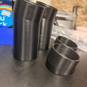 May include: Several black 3D-printed plastic tubes. Two are angled and larger, while two are smaller and ring-shaped. The items are displayed on a dark surface.