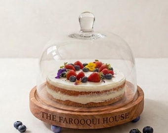May include: A round wooden cake stand with a glass dome lid. The base is engraved with the text THE FAROOQUI HOUSE. Inside the dome sits a two-layer cake topped with white frosting, fresh strawberries, blueberries, and yellow flower petals.