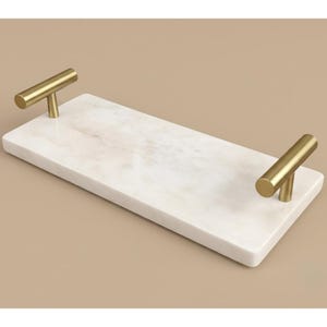 May include: A rectangular white marble serving tray featuring a polished surface with subtle grey veining. The tray is equipped with two cylindrical gold-toned metal handles attached to each of the shorter ends, providing a secure grip for carrying.