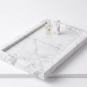 May include: A rectangular white marble tray with grey veining featuring raised edges. Resting on the surface of the tray are two small glass bottles, one square with a silver cap and one cylindrical with a silver cap and a pink label.