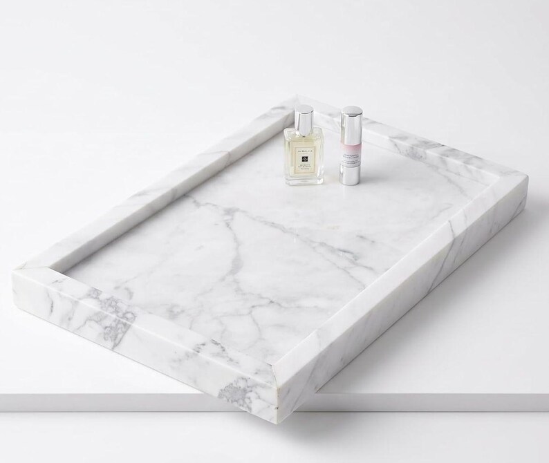 May include: A rectangular white marble tray with grey veining featuring raised edges. Resting on the surface of the tray are two small glass bottles, one square with a silver cap and one cylindrical with a silver cap and a pink label.