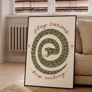 May include: A framed rectangular wall art print on a cream background featuring a coiled green snake with an open mouth and red tongue. The text "stay curious" is written in red above the snake, and "keep evolving" is written in red below.