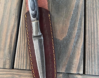 Può includere: Un coltello a lama fissa con una lama appuntita in acciaio damasco caratterizzata da un motivo ondulato. Il manico è realizzato in legno grigio scuro e nero con dettagli in metallo. Il coltello è parzialmente inserito in un fodero in pelle marrone.