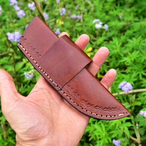 Puede incluir: Una funda de cuchillo de cuero marrón con costuras decorativas y cierre de solapa. La funda está diseñada para guardar un cuchillo. El fondo muestra follaje verde y flores moradas.