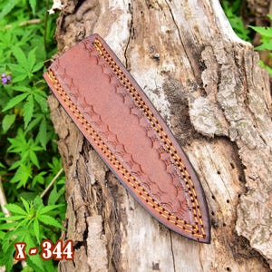 May include: Brown leather knife sheath with decorative stitching and embossed design. The sheath is shown on a weathered wooden surface with green foliage in the background. The text "X-344" is visible in the lower left corner.
