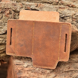 May include: A brown leather holster with yellow stitching. The holster has two vertical slots and a flap. The leather has a distressed appearance. The item is displayed on a piece of wood.