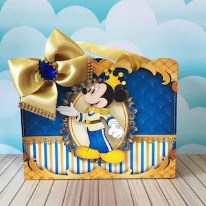 May include: A decorative box featuring a cartoon image of Mickey Mouse in a prince outfit. The box has a blue and gold color scheme, with a large gold bow and a blue jewel. The box has a striped pattern at the bottom.