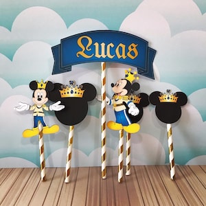 May include: Cake toppers featuring Mickey Mouse in royal attire, with a banner that reads "Lucas". The toppers include Mickey Mouse figures, Mickey Mouse head silhouettes with crowns, and gold and white striped sticks.