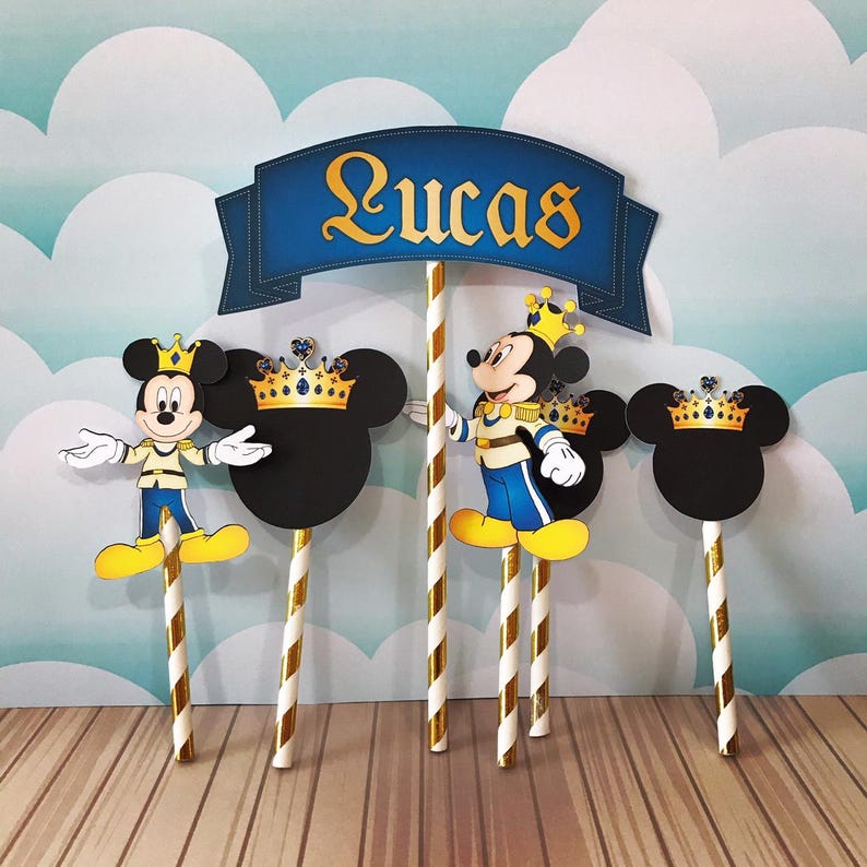 May include: Cake toppers featuring Mickey Mouse in royal attire, with a banner that reads "Lucas". The toppers include Mickey Mouse figures, Mickey Mouse head silhouettes with crowns, and gold and white striped sticks.