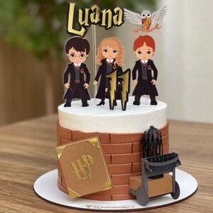 Könnte beinhalten: Ein dekorierter Kuchen mit Cartoonfiguren von drei Charakteren in schwarzen Roben, mit dem Text "Luana" oben. Der Kuchen hat ein braunes Ziegeldesign an den Seiten, mit einem goldenen Buch und einem Wagen mit Rädern.