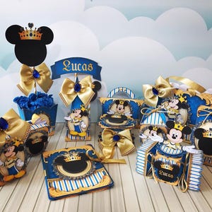 May include: A collection of Mickey Mouse-themed party decorations in blue, gold, and white. The decorations include boxes, bows, and cutouts, some with the name "Lucas". The Mickey Mouse silhouette is featured on several items, with a crown.