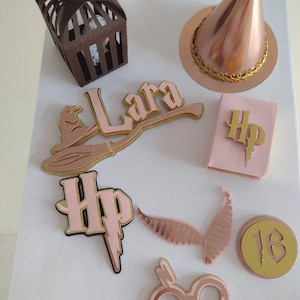 Op de afbeelding: Een verzameling roze gouden en roze Harry Potter-themafeestdecoraties. Inclusief een heksenhoed, een kooi, een bezem, de naam "Lara", de letters "HP", een boek, een bril, vleugels en het nummer 16. Op een wit oppervlak.