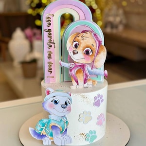 Könnte beinhalten: Eine weiße Torte, verziert mit Zeichentrickfiguren und einem Regenbogenbogen. Die Torte zeigt Skye und Everest von Paw Patrol, mit Pfotenabdrücken und goldenen Glitzerakzenten. Der Text auf dem Bogen lautet "Essa garota vai voar!"