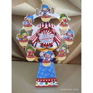 Op de afbeelding: Een kleurrijke, decoratieve verjaardagsversiering in de vorm van een reuzenrad met cartoonclowns. De clowns staan in verschillende poses en de versiering heeft een blauw, rood en wit kleurenschema. De tekst "Enrico 3 anos" is zichtbaar.