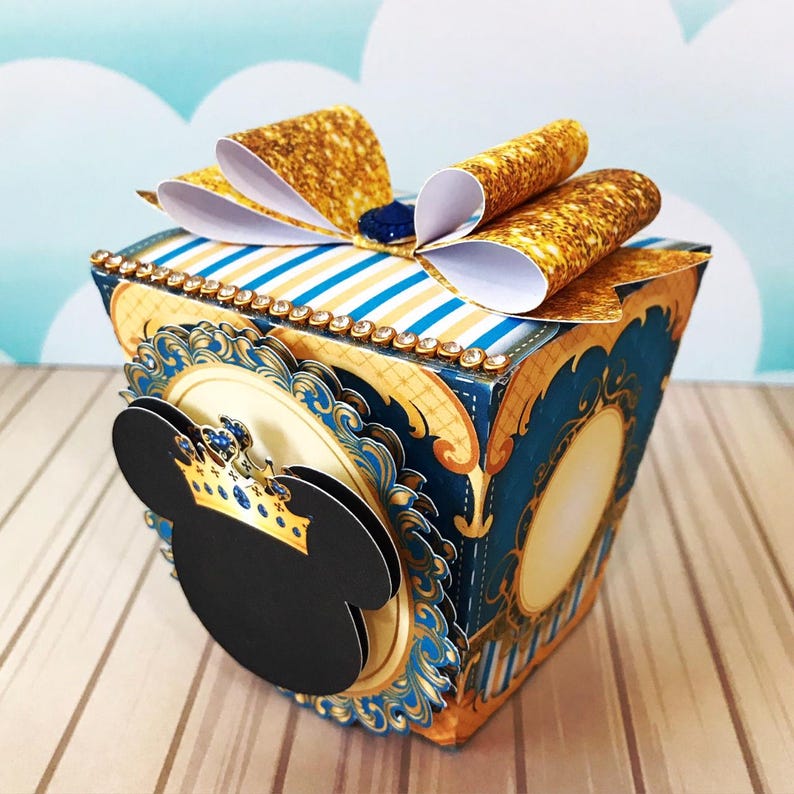 May include: A decorative box with a gold bow and a Mickey Mouse silhouette. The box is blue with gold accents and a striped top. The Mickey Mouse silhouette has a gold crown with blue jewels.