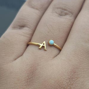 Può includere: Un anello aperto color oro con una lettera A maiuscola su un'estremità e una piccola pietra rotonda azzurra sull'altra. La fascia è sottile e metallica, progettata per essere indossata attorno al dito con le due estremità rivolte l'una verso l'altra.