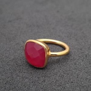 May include: A gold-toned ring featuring a single, faceted, cushion-cut dark pink gemstone set in a smooth metal bezel. The band is plain and rounded, showcasing a polished finish that contrasts with the textured surface of the stone setting.