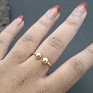 Op de afbeelding: Een goudkleurige open ring met twee gepolijste bolvormige kralen aan elk uiteinde van de band. De ring is ontworpen met een opening tussen de twee bollen, waardoor een eenvoudige, doorlopende metalen lus ontstaat die de vinger glad omsluit.