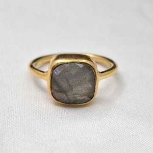 May include: A gold ring featuring a square-cut, faceted gray gemstone with subtle iridescent undertones. The stone is set in a smooth, polished gold bezel, connected to a thin, rounded gold band that completes the jewelry piece.