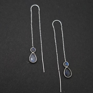 Puede incluir: Un par de pendientes de hilo de plata con un diseño de cadena fina. Cada pendiente está decorado con una pequeña piedra redonda y una piedra más grande en forma de lágrima, ambas facetadas y con un sutil brillo azul iridiscente.