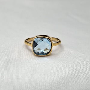 May include: A gold ring featuring a square-cut, faceted light blue gemstone set in a polished gold band. The stone has a checkerboard pattern on its surface, reflecting light with a clear, transparent blue hue against the gold setting.