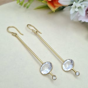 Pode incluir: Um par de brincos compridos dourados com uma barra vertical fina presa a um gancho. Na base de cada barra existe uma pedra grande oval transparente facetada, com uma pedra redonda mais pequena fixada por baixo num engaste dourado.