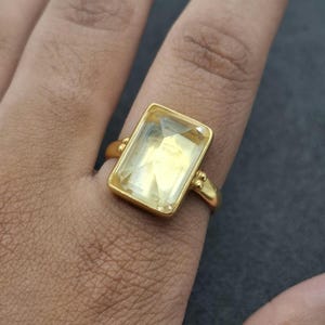 Könnte beinhalten: Ein goldener Ring mit einem großen, rechteckigen, facettierten blassgelben Edelstein in einer glatten Metallfassung. Die Ringschiene ist schlicht und poliert, mit kleinen dekorativen Metallkugeln an der Verbindung zur Steinfassung.