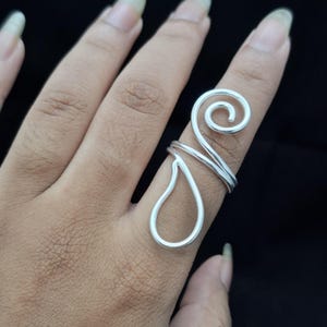 May include: A silver-colored ring featuring a continuous wire design that forms a spiral at the top and a teardrop-shaped loop extending downward along the finger. The metal has a smooth, polished finish and a simple, open-ended construction.