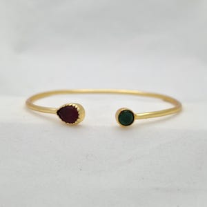 Puede incluir: Un brazalete abierto de color dorado. Un extremo presenta una piedra preciosa roja oscura en forma de lágrima, el otro una piedra preciosa verde oscura redonda. El brazalete está sobre un fondo blanco.