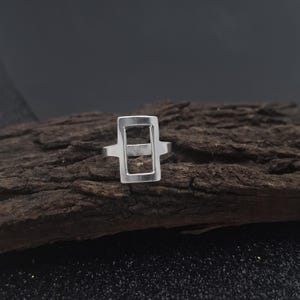 May include: A silver-colored ring with a rectangular design. The ring features a hollow, open rectangle with a horizontal bar across the middle. The ring is set against a dark, textured wooden surface.