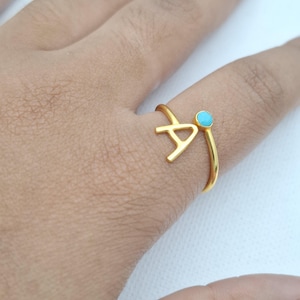 Peut inclure: Une bague dorée ornée de la lettre "A" et d'une petite pierre turquoise ronde. La bague est portée sur un doigt, mettant en valeur son design délicat et le contraste de couleurs vives.