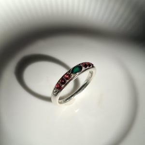 May include: A silver ring featuring a central marquise-cut green gemstone flanked by three small round red gemstones on each side. The band is polished silver, and the stones are set in a row along the top portion of the circular metal band.