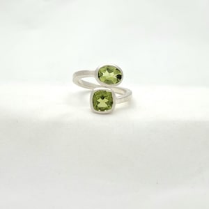 May include: A silver ring featuring two light green peridot gemstones. One gemstone is oval-shaped, and the other is square-shaped. The ring's band wraps around the finger, creating a unique and modern design. The gemstones are set in a bezel setting.