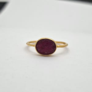 May include: A gold ring with a large, oval, deep red gemstone. The ring's band is thin and gold. The gemstone is the focal point, set in a simple, elegant design. The ring is displayed on a white surface.